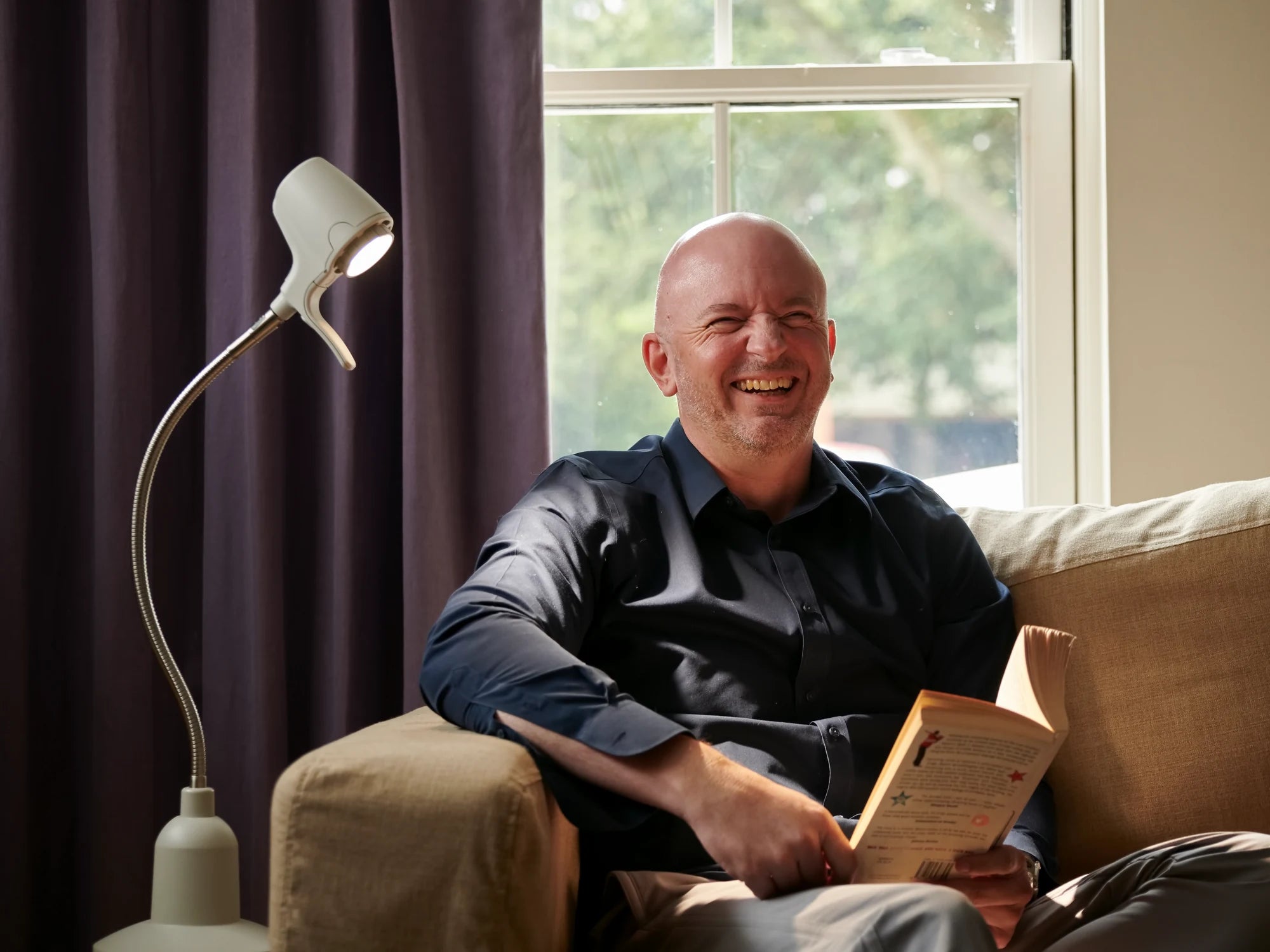 A man laughing over his book on his sofa, beside an HD Original Table Light in Heritage White