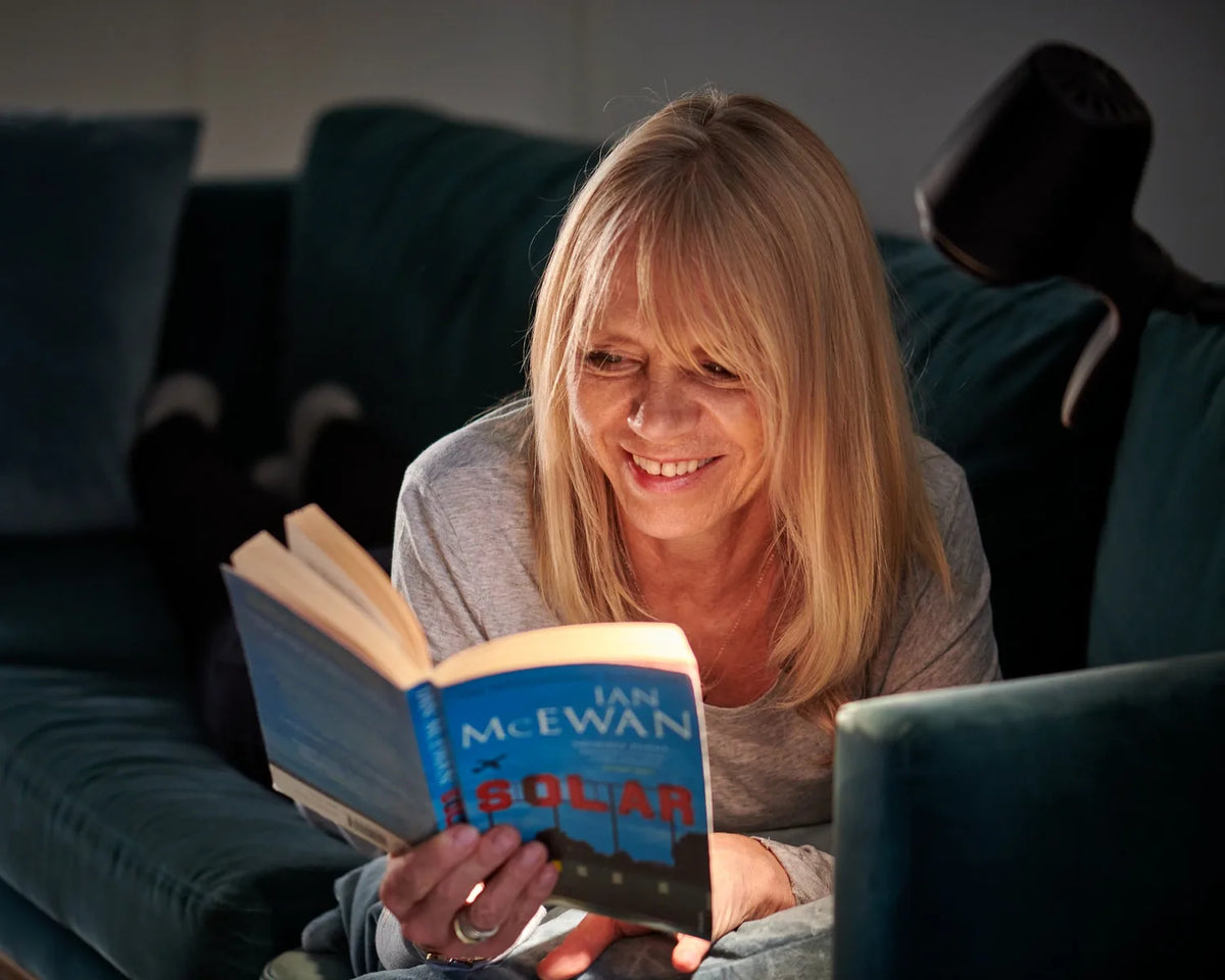 A woman lying on her sofa at night, smiling over her book under an HD Original Table Light