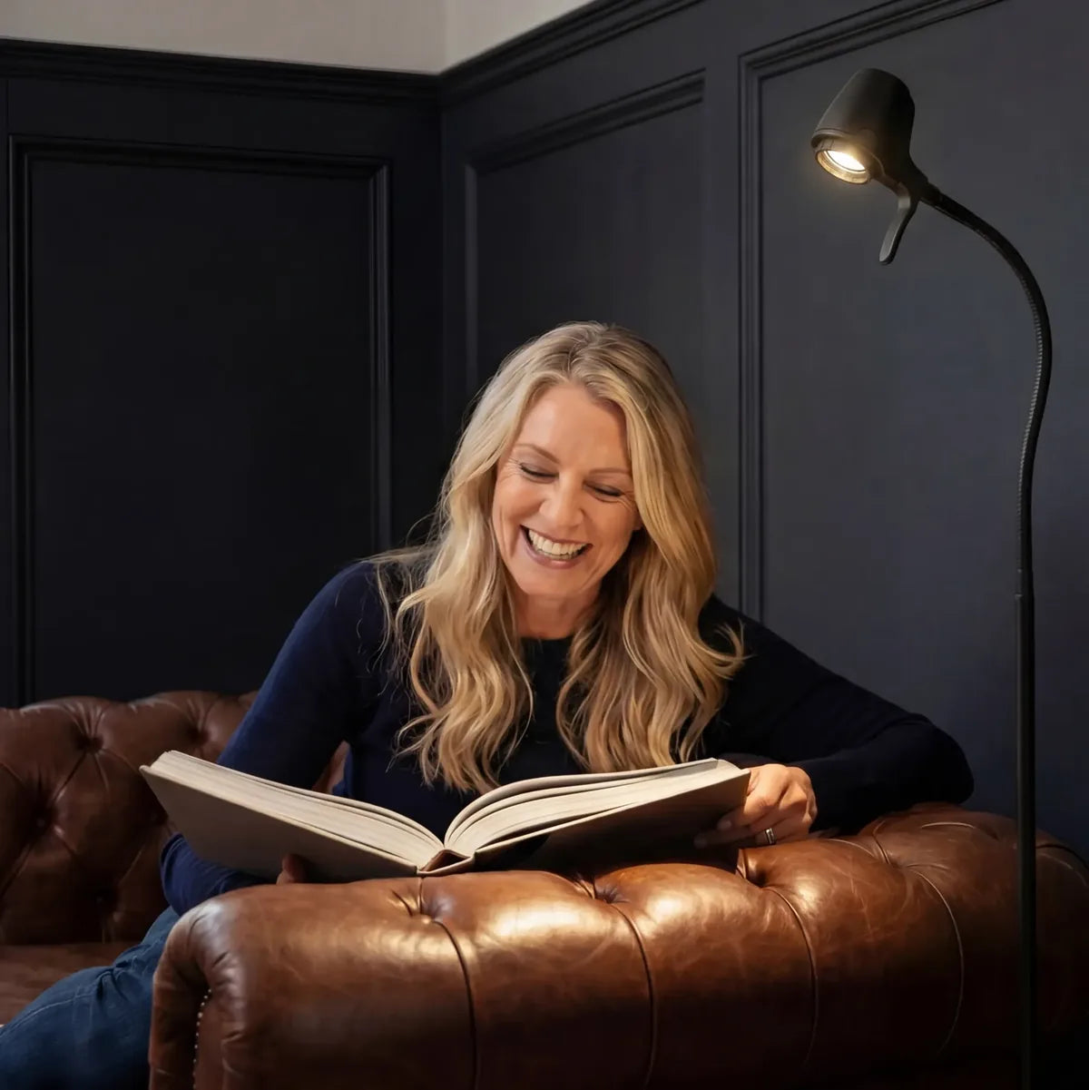 A woman laughing as she reads a large book on a leather sofa, beside a Serious floor light