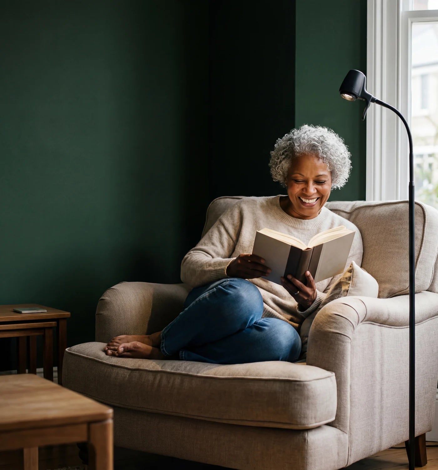 A woman laughing over her book in a cream armchair beside an HD Pro Floor Light