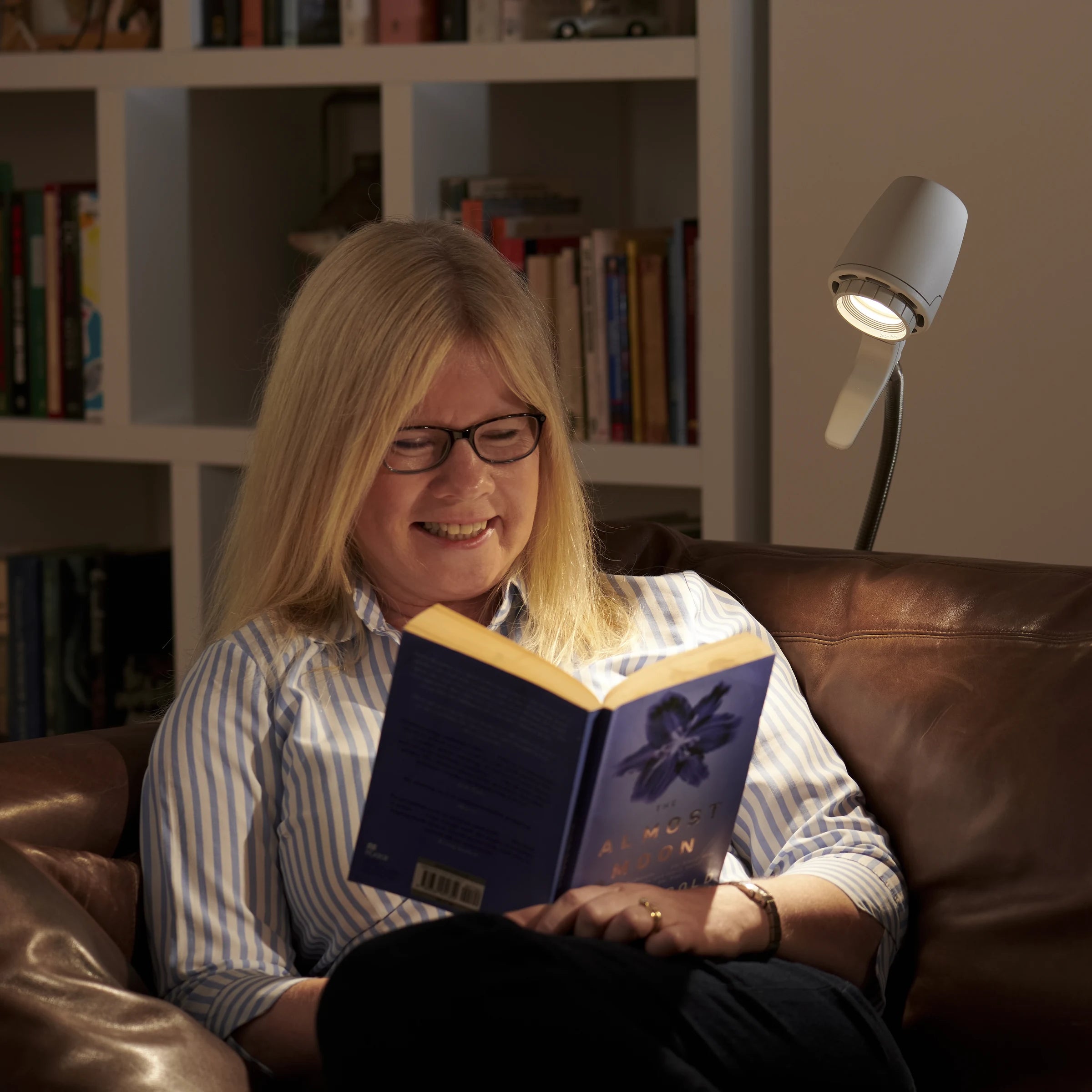 A woman smiling over her book on a leather sofa, lit by an HD Original Floor Light in Heritage White