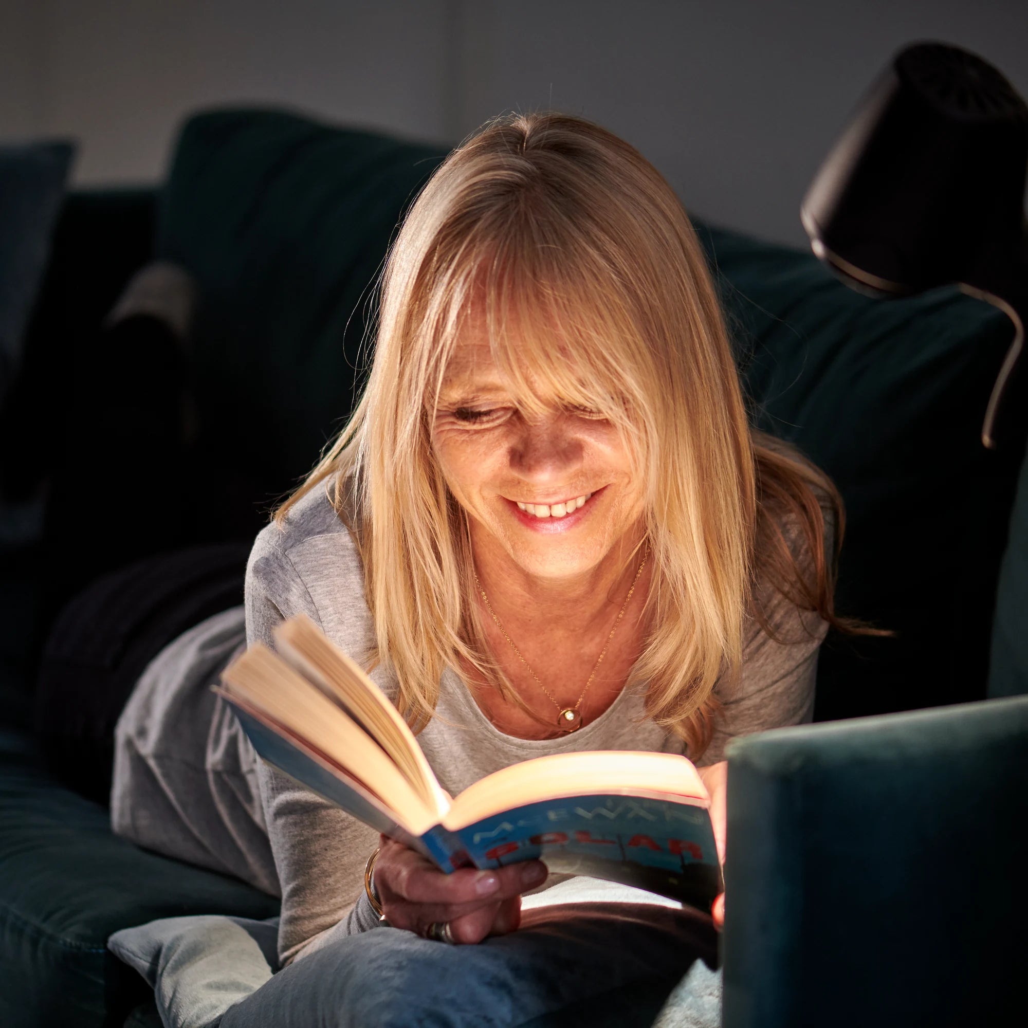 A woman smiling over her book on her sofa by the light of an HD Original Floor Light