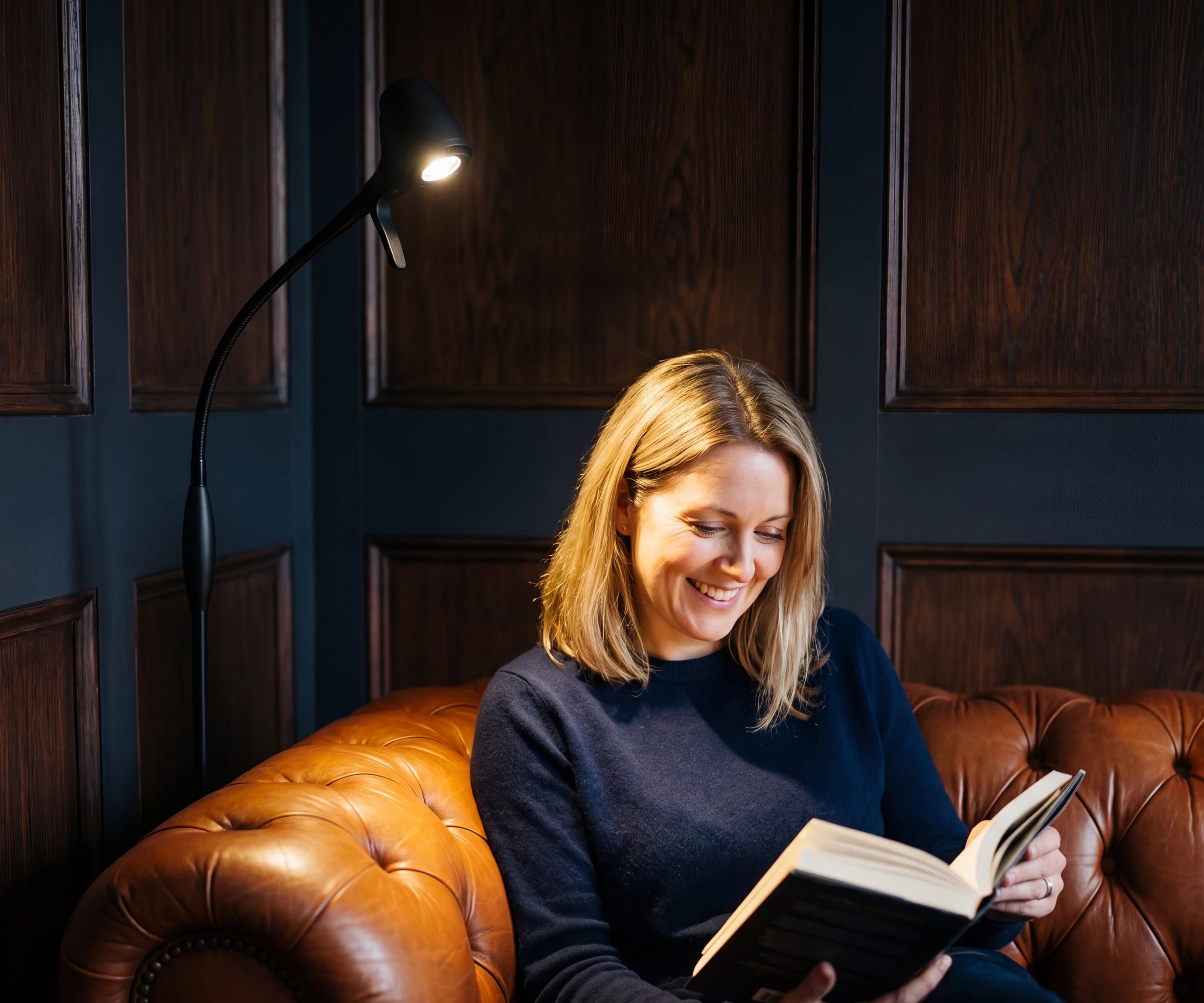 A woman laughing over her book on a leather chesterfield in the evening, lit by an HD Original Floor Light