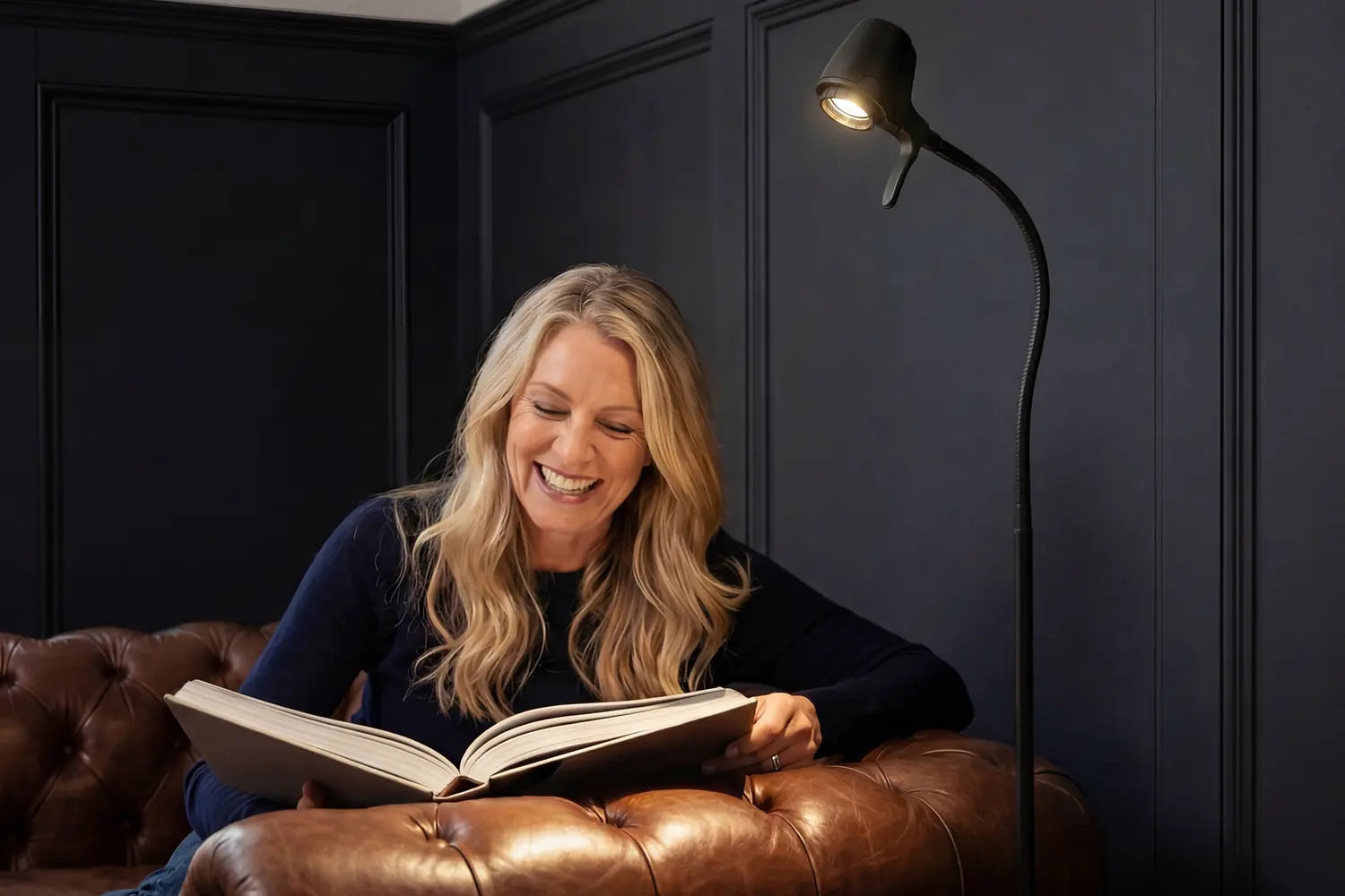 A woman laughing over her book on a leather sofa beside a Serious floor light