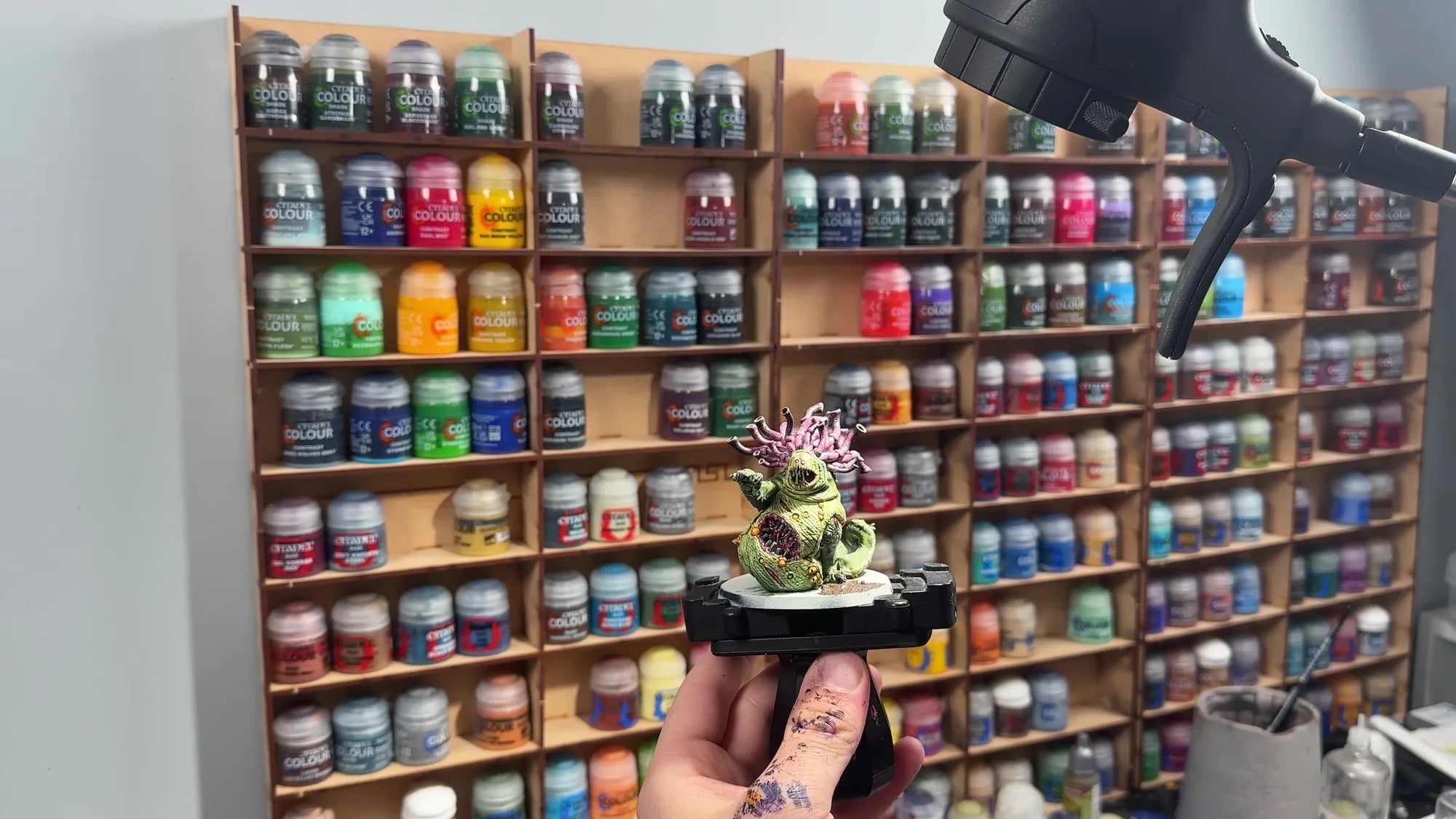 A painted miniature held up in front of shelves of paint pots, under the head of a Serious Light