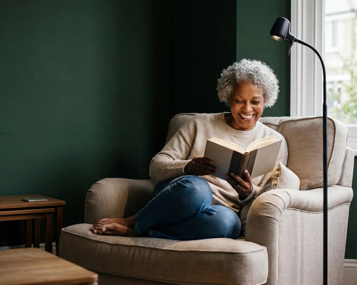 A woman laughing over her book in an armchair beside an HD Pro Floor Light